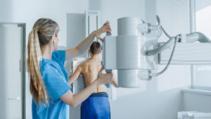 In The Hospital, Man Standing Face Against The Wall While Medical Technician Adjusts X Ray Machine For Scanning. Scanning For Fractures, Broken Limbs, Chest, Cancer Or Tumor. Modern Hospital With Technologically Advanced Medical Equipment.